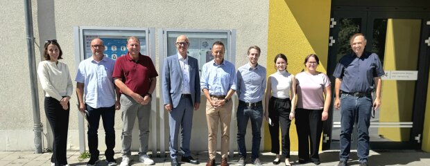 Gruppenfoto vor dem Eingangsbereich des ANKER Oberpfalz. Acht Personen stehen nebeneinander in der Sonne. In der Mitte stehen Regierungspräsident Walter Jonas und der Integrationsbeauftragte der Bayerischen Staatsregierung, MdL Karl Straub, umgeben von Mitarbeitenden des ANKER. Rechts im Hintergrund ist eine gelb gestrichene Gebäudeecke mit Eingangstür zu sehen. Regierungspräsident Walter Jonas (4. v. l.) und der Integrationsbeauftragte MdL Karl Straub (5. v. l.) mit Mitarbeitenden des ANKER Oberpfalz.